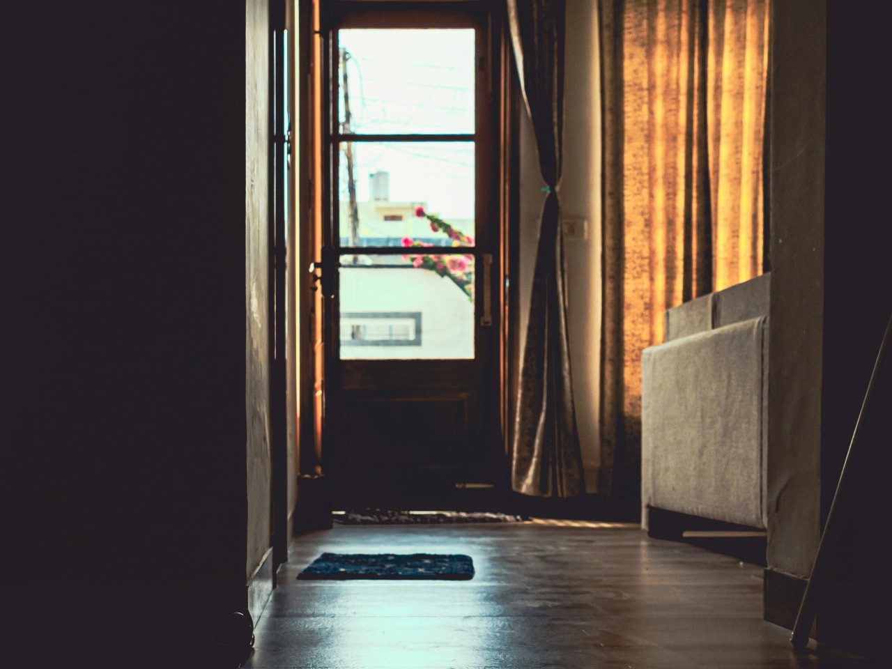 A dark hallway with light reflecting off the floor, leading toward a bright open doorway framed by warm curtains, representing practicing beginning from want, not from duty, fear, or performance.